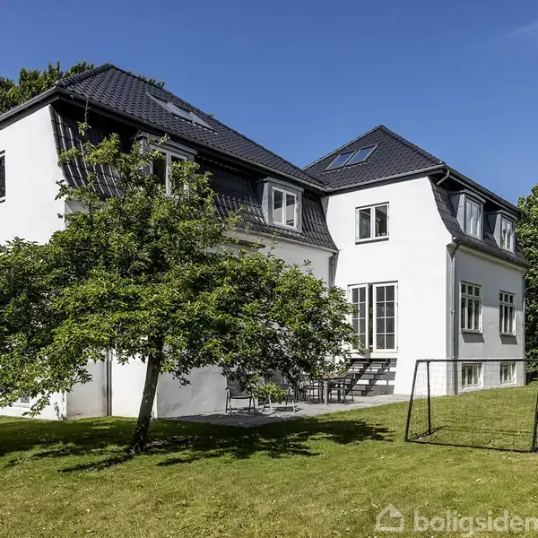 Hvidt, to-etagers hus med sort tag står blandt træer i en grøn have. Sollys skinner på facaden. Der er en fodboldmålramme på plænen.