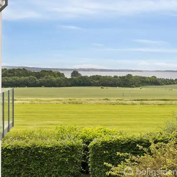 Bygning med glasaltan står på venstre side, og udsigten viser en grøn mark omkranset af træer med en fjord i baggrunden under en klar blå himmel.