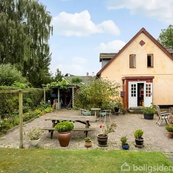 Et klassisk hus med skråtag står i en brostensbelagt gårdhave fyldt med krukker, træmøbler og grønne planter. En stor grøn have og træer omkranser området.