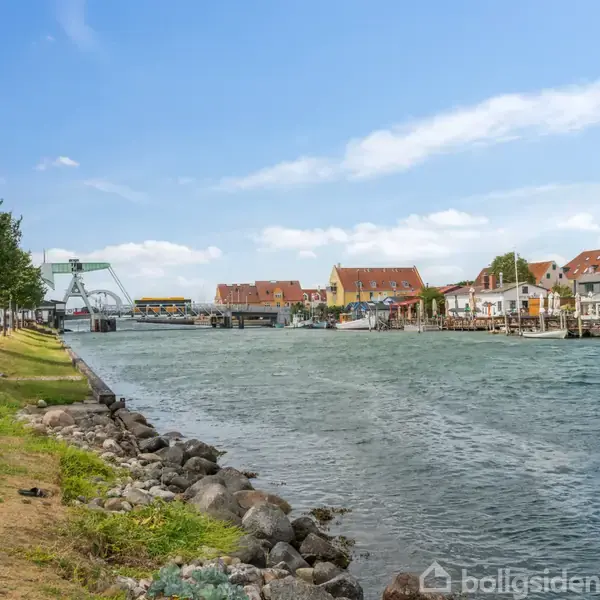 Flod løber gennem by med rødt tage og træer langs bredden; en bro ses i horisonten under en klar blå himmel.
