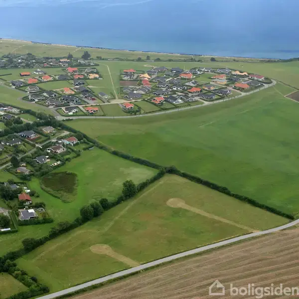 Boligområde med spredte huse omkranset af veje og grønne marker. Bebyggelsen ligger i et åbent landskab nær en kystlinje. Forskellige hustagefarver ses tydeligt.