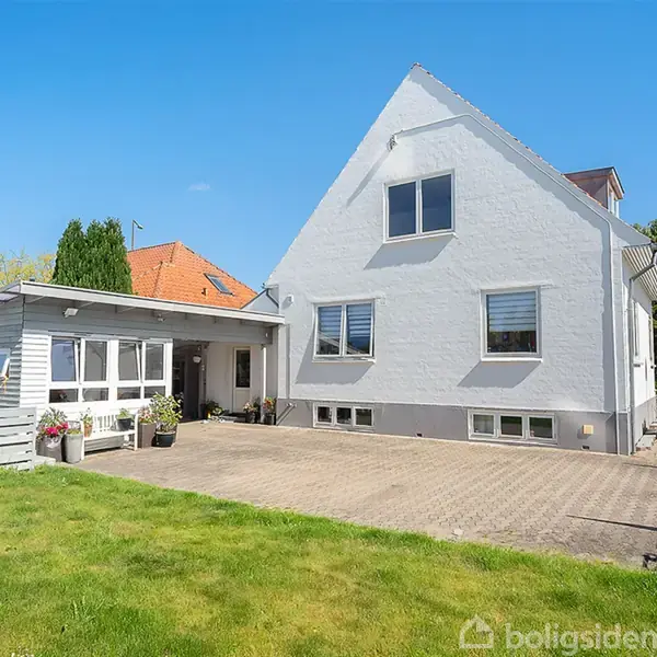 Hvidt hus med gavltag står i en solrig have med grøn græsplæne. Tilknyttet carport med blomsterkrukker. Klar blå himmel dominerer baggrunden.