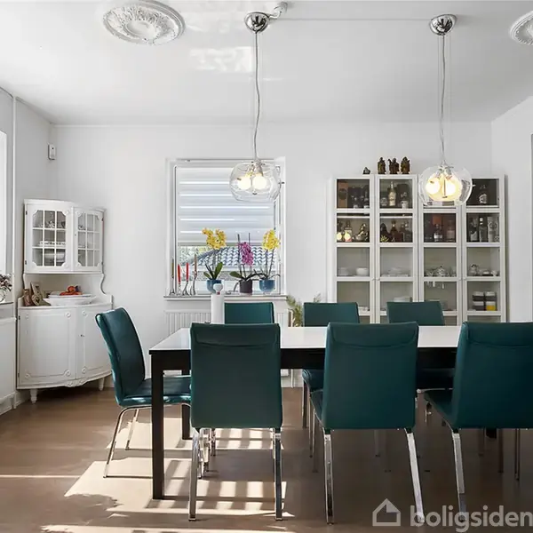 Spisebord med blå stole står i et lyst rum. To lamper hænger over bordet. Skabe med glaslåger og udsmykkede loftsdetaljer ses i rummet.