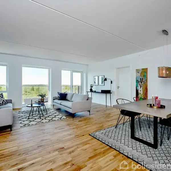 To sofaer står i en lys stue med trægulv og store vinduer. Et spisebord med sorte stole står i hjørnet. Væggene er dekoreret med malerier.