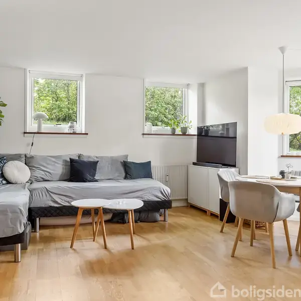 En stue med en stor hjørnesofa udstyret med puder. Ved siden af står et fjernsyn på et møbel. To runde borde foran sofaen. Et spisebord i hjørnet med stole. Tre vinduer slipper lys ind og tilbyder udsigt til grønne træer. Plante hænger ved sofaen; en lampe hænger over spisebordet. Trægulv skaber en varm atmosfære.