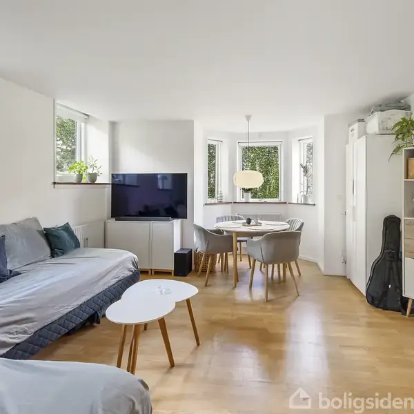 En lys stue med trægulv har en grå sofa, et fladskærms-tv og et spisebord med stole. Rummet er omgivet af grønne planter og store vinduer.