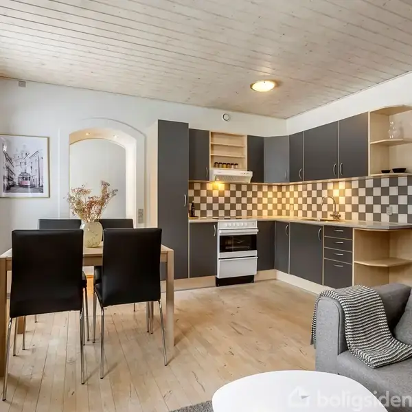 Et moderne køkken-alrum med lyse trægulve og træloft. Køkkenet har grå skabe og en skakternet backsplash. Der står et spisebord med fire stole. En sofa ses i forgrunden.