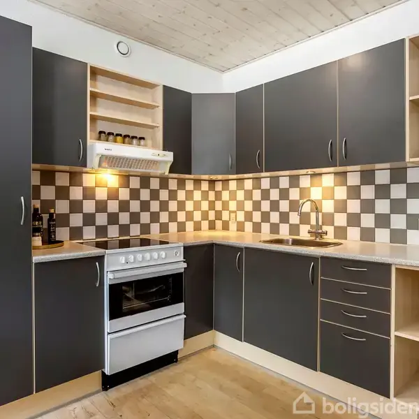 Et køkken med grå skabe omkranser en hvid komfur og vask. Skakternet vægflisebaggrund skaber kontrast i rummet. Hylder med krydderier og håndklæde tilføjer praktiske detaljer.