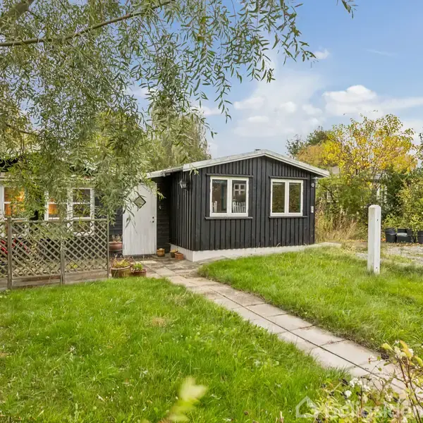 Et sort træhus med hvide karme står stille i en frodig have. En flisebelagt sti leder til hoveddøren, omkranset af grønne græsplæner og buske.