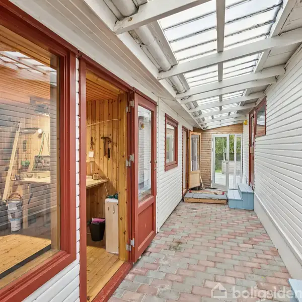 En lys gang med glastag og røde dørkarme leder til flere rum. På venstre side ses et værksted fyldt med værktøj. Gangens gulv er belagt med fliser.