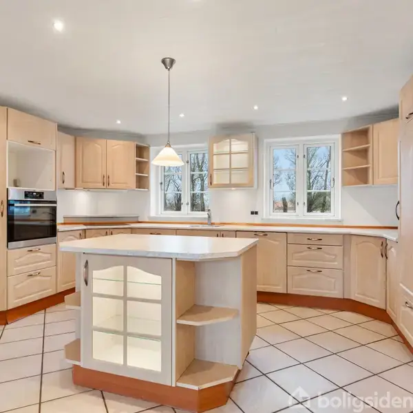 Et moderne køkken med lyse træskabe omkranser et centralt kogeø. Ovn indbygget i væggen. Store vinduer bag kogeøen lader naturligt lys strømme ind. fliser på gulvet.