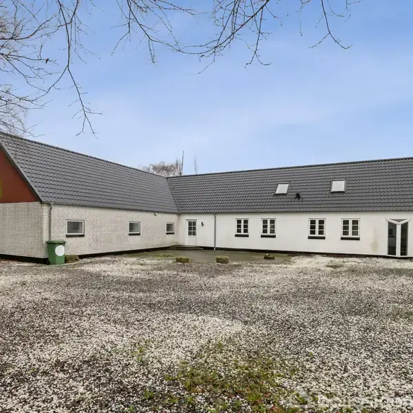 En hvid bygning med mørkt tag står på en grusbelagt gårdsplads, omgivet af træer uden blade. Bygningen har flere vinduer og en grøn affaldscontainer ved siden af.