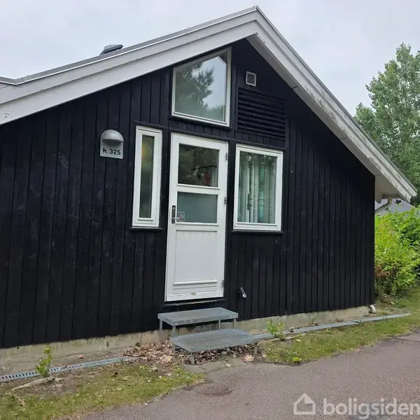 Sortmalet hus med hvid dør og vinduer, der vender ud mod en sti omgivet af grønt løv. Et skilt ved døren viser "K 325".