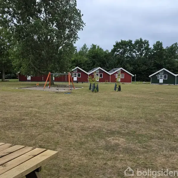 Legeplads med gynger står stille på en åben græsplæne, omgivet af små, farverige hytter og grønne træer i baggrunden.