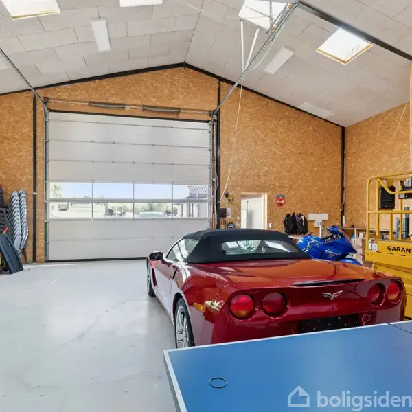 En rød sportsvogn står parkeret i en stor garage med højt til loftet. Ved siden af er der en grøn hvid varevogn og en gul lift med teksten "GARANT UDLEJNING". En blå bordtennisbord ses i forgrunden.
