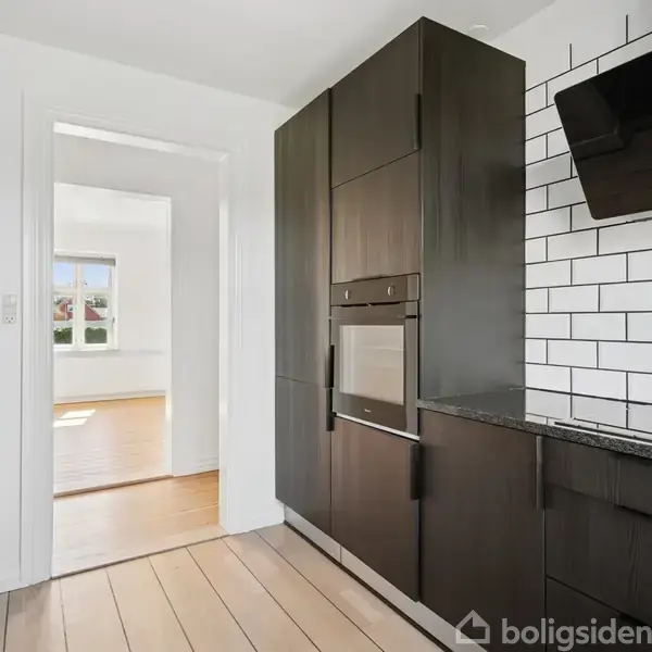 Et moderne køkken med mørke skabe og integreret ovn står i fokus. En hvid flisevæg tilføjer kontrast. I baggrunden ses en åben dør ind til et lyst rum.