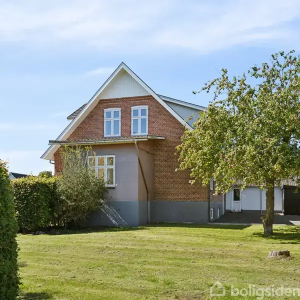 Et rødt murstenshus med hvidrammede vinduer står i en velplejet have med en græsplæne og et stort træ. Huset har en grå sokkel og et gavltag.