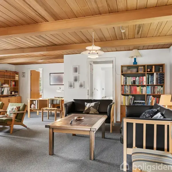 Stue med trægulv og træloft, møbleret med sorte lædersofaer og træstole omkring et sofabord. Bogreol ved væggen. Vinduer med udsigt til grønne omgivelser. Hyggelig atmosfære.