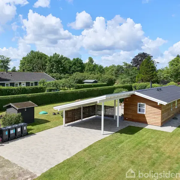 Huset står fredeligt omgivet af en grøn have og hække. Det er et træhus med fladt tag og en overdækket terrasse. En flisebelagt indkørsel fører op til huset.
