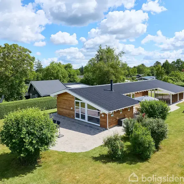 Et moderne hus med træfacade ligger midt i en velplejet have med græsplæne og buske. Huset har store vinduer, sort tag og er omgivet af træer under en blå himmel med skyer.