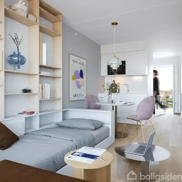 En grå sofa fungerer som en seng i en moderne stue med hylder fyldt med bøger og dekorationer. Et rundt spisebord med stole står i nærheden under en pendellampe. En plakat på væggen viser teksten "NON ARTE GRAFICA." En person bevæger sig i baggrunden nær altandøren.