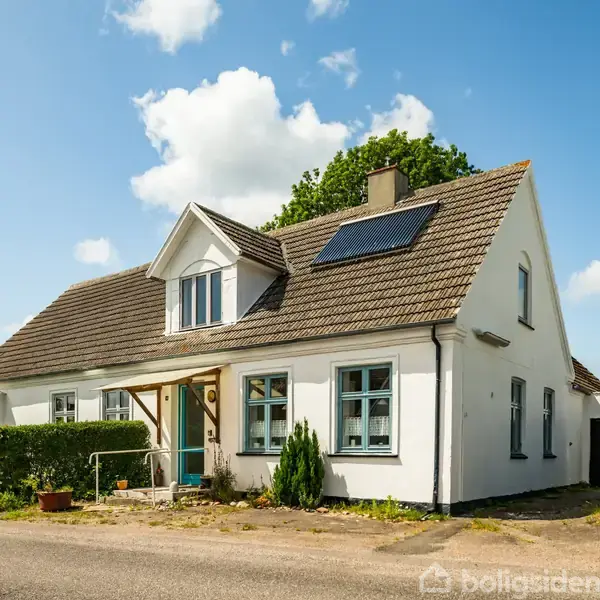 Et hvidt hus med skråt tag og en solpanel, står ved en vej. Der er en grusvej og en hæk i baggrunden under en blå himmel med skyer.