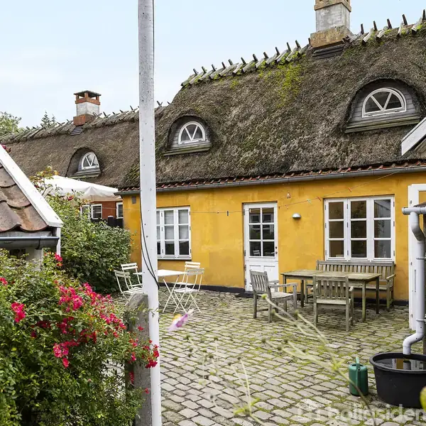 Gult bindingsværkshus med stråtag og runde vinduer, omkranset af blomstrende buske. På brostensbelagt terrasse står hvide stole og haveborde, omgivet af frodig vegetation og blå himmel.