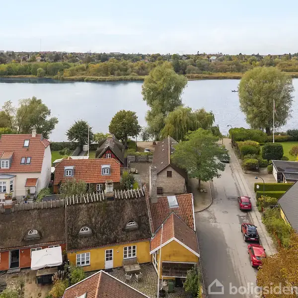 Traditionelle stråtækte huse står langs en fredelig vej med parkerede biler. I baggrunden breder en sø sig, omgivet af træer i efterårsfarver, hvilket skaber en naturskøn atmosfære.
