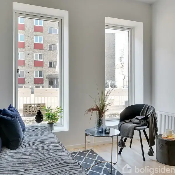 En lille, moderne stue med en grå sofa fyldt med blå puder står langs væggen. Ved siden af er et rundt bord og en sort stol, alt oplyst af naturligt lys fra store vinduer.