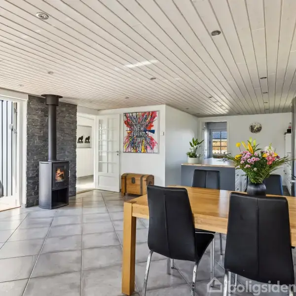 Brændeovn opvarmer stue med flisegulv og træloft. Bord med blomster og seks stole ved køkken. Farverigt maleri på væg. Glasdøre fører til terrassen. Køleskab nær køkkenet.