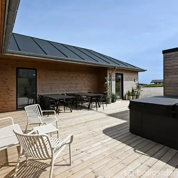 Terrasseområde med hvide stole og et sort spabad, bord og stole under overdækning. Omgivet af træbeklædte bygningsfacader, med åben udsigt over landskab.