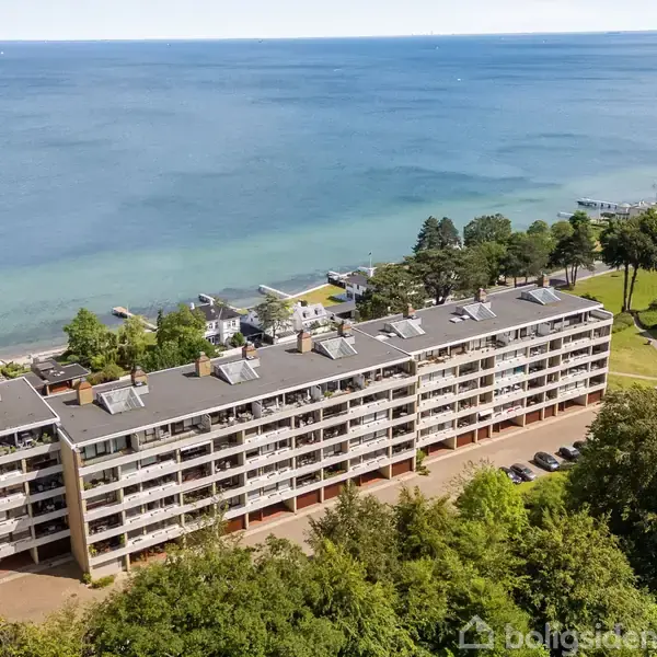 Et længere hvidt etagebyggeri strækker sig langs kysten, omgivet af træer og tæt på havet. Bygningen har fem etager og balkon på hver etage.