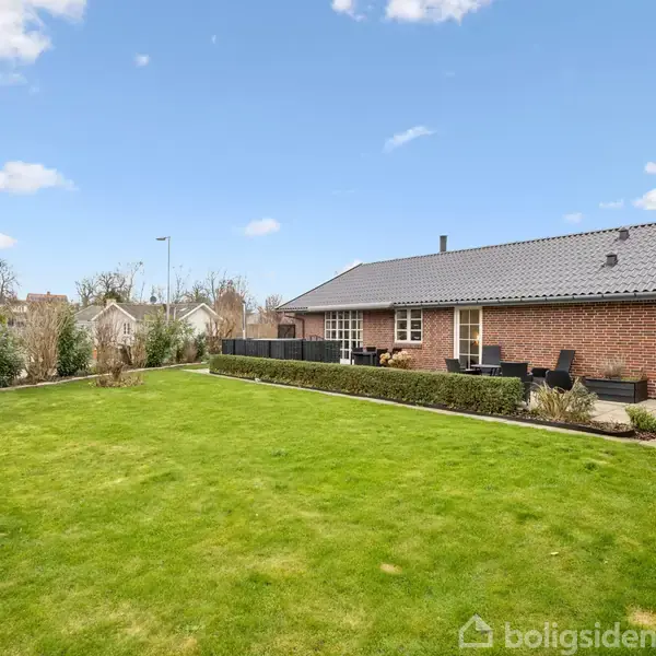 En rød murstensbygning står stille, omgivet af en velplejet grøn græsplæne og flisebelagt terrasse. I baggrunden ses buske og en klar blå himmel med få skyer.