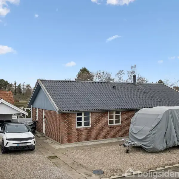 Rødt murstenshus med sort tag står på en grusbelagt gårdsplads. En bil er parkeret ved siden af et overdækket køretøj. Omkring er der buske og træer under en blå himmel.