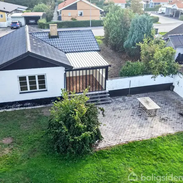 Et hus med sort tag har en terrasse med trapper ned til en brolagt gårdhave. I gårdhaven står et langt bord. Omkring er grønne buske og træer.