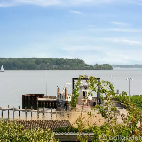 Færge ventes ved en havn, omgivet af grøn vegetation og en molen. I baggrunden ses en søjlerække, en sejlbåd på vandet og en skovklædt kystlinje under en lyseblå himmel.