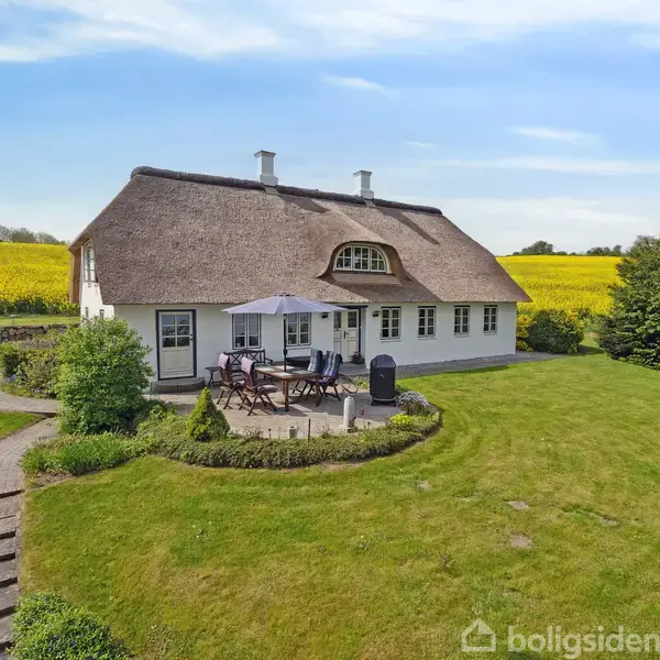 Et traditionelt bondehus med stråtag står i en frodig have, omgivet af marker. Huset har en lille terrasse med borde og stole, ideelt til udendørs afslapning.