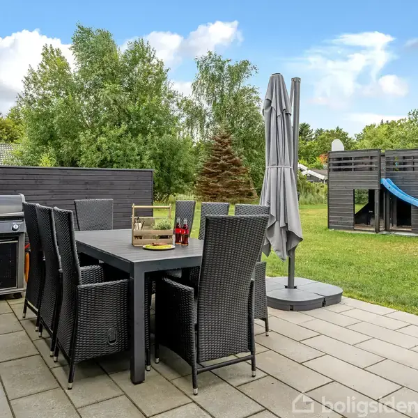 Terrasseområde med spisebord og stole står ved grill. Parasol giver skygge. I baggrunden ses en græsplæne med legehus, rutsjebane og gynger. Træer omkranser området.