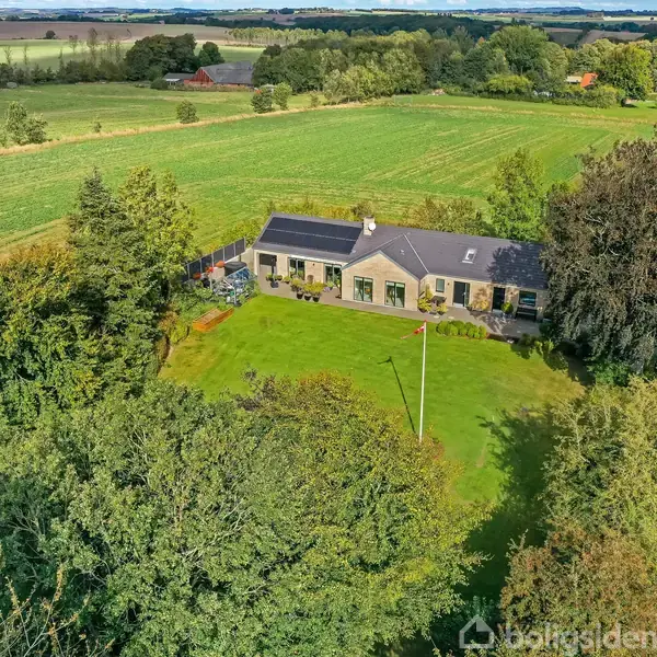 Hus ligger i landligt område omgivet af træer og græsplæne. Grønne marker strækker sig ud i baggrunden. Veje omkranser ejendommen på højre side.