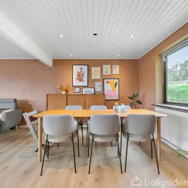 Spisebord med seks grå stole i en stue. Bordet er dækket med vandflasker og glas. Omgivet af kunstplakater på væggen og et vindue med udsigt til haven.