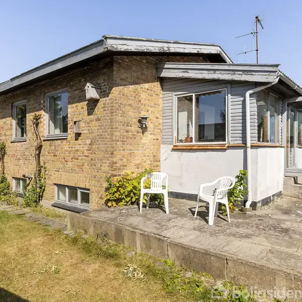 Et murstenshus med fladt tag står stille i en solrig have, omgivet af græs og træer. To hvide plastikstole står på en flisebelagt terrasse foran huset.