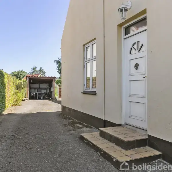 Hvid dør med glas og lanterne hænger på husmur; foran trin med fliser. Omgivet af hæk, grusvej fører til carport i baggrunden. Ingen tekst fundet.