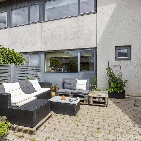 En udendørs terrasse har moderne sort, rattanmøbler med hvide puder. Terrassen er omgivet af planter og har en stakitthegn til venstre. Bygningens facade er lys med store vinduer.