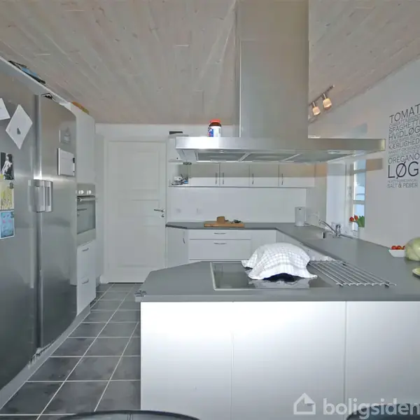 Køkken med rustfrit stållig køleskab, integreret komfur på ø. Moderne hvide skabe. Opslagstavle med små papirer. Tekst på væg: "Tomat, løg, bacon." Flisegulv, loft med spots.