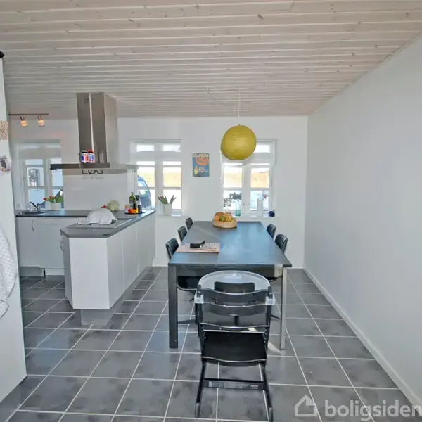 Sort spisebord med stole står midt i rum. Køkken ses i baggrunden med hvide skabe. Vægur hænger på hvid væg til højre. Gul lampe hænger fra loftet.