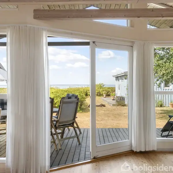 Vinduer med hvide gardiner åbner ud til en terrasse med stole og bord. Sollys strømmer ind, og i baggrunden ses en have og en lys himmel.