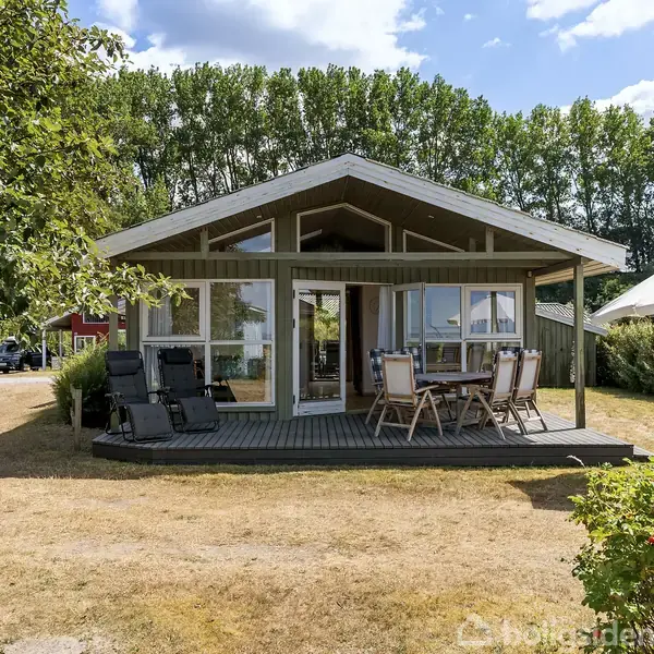Et sommerhus i træ står på en tør græsplæne, omgivet af buske og træer. Foran huset er der en terrasse med havemøbler og en parasol.