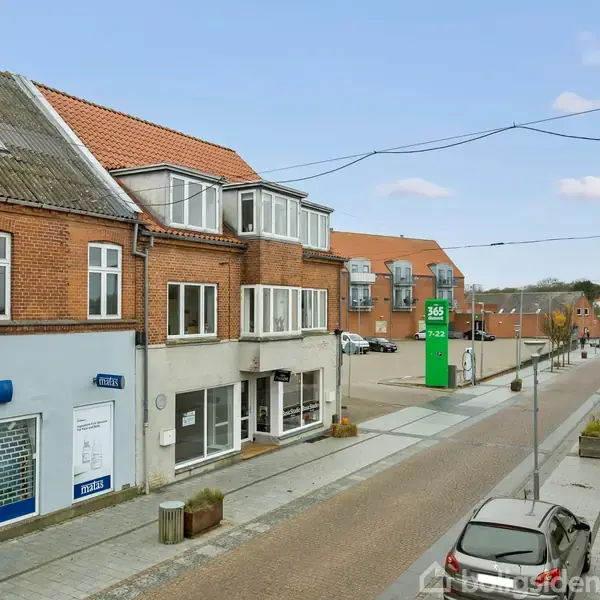 Et gadebillede viser røde murstensbygninger med butikker, herunder en "Matas" butik, langs en stille vej med parkerede biler. En grøn 365-skiltepæl står ved et moderne hjørnekompleks.
