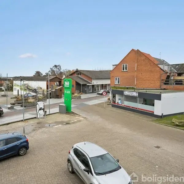 En grå bil holder stille på en parkeringsplads ved siden af en vej. En grøn tankstation med teksten "365 92 R2" og en bygning ses i baggrunden.