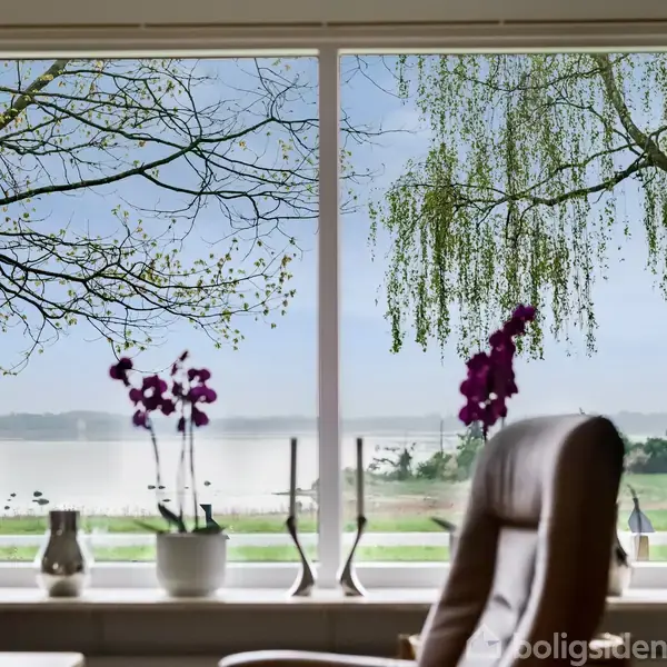 Stol står foran et stort vindue, omgivet af blomster og planter, med udsigt over en sø og træer i baggrunden. Rolig atmosfære med naturskøn udsigt gennem vinduet.